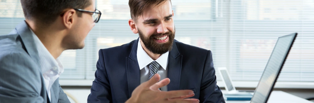 Two businessmen sitting at a desk talking by a computer
